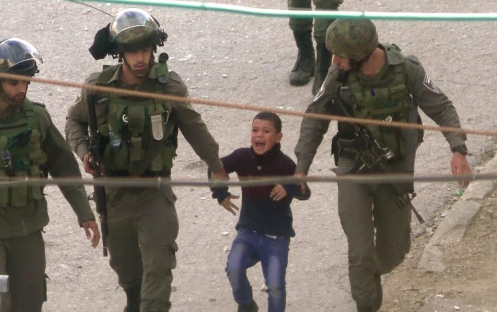 Stillbilde fra filmen, som viser en ung gutt føres bortover av tre voksne, bevæpnede soldater. Guttens munn er åpen, som om han roper noe.